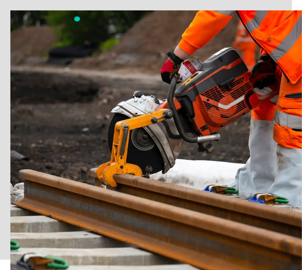 Worker in orange safety gear cutting a steel rail with a concrete saw at a construction site.