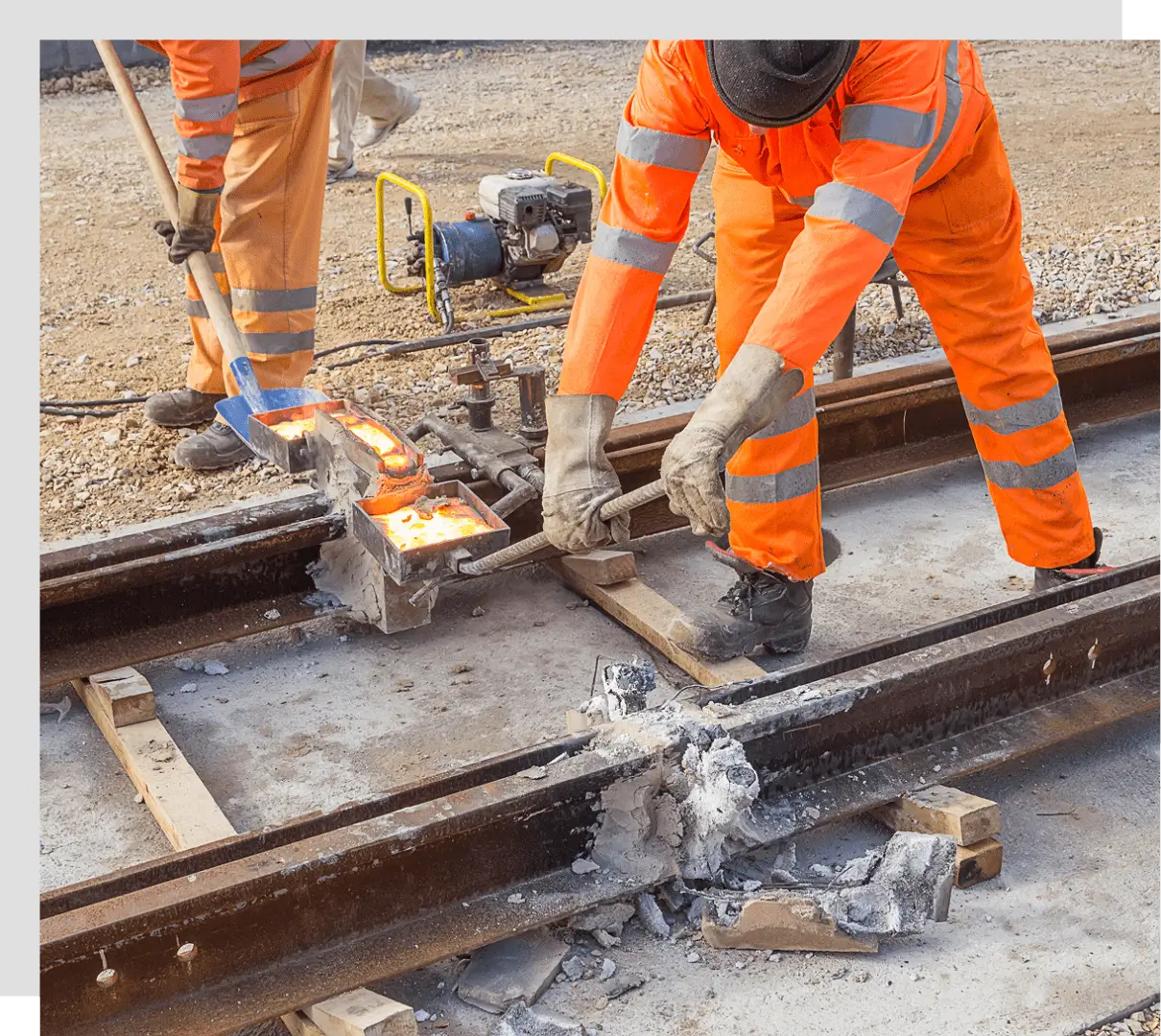 Workers in orange uniforms repair railway tracks using equipment and tools, surrounded by pieces of concrete and debris.