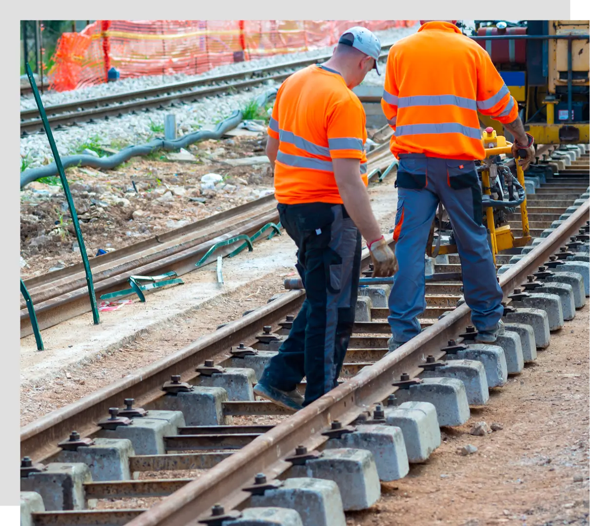 Two workers in orange safety gear inspect a railway track under construction, surrounded by gravel and construction materials.